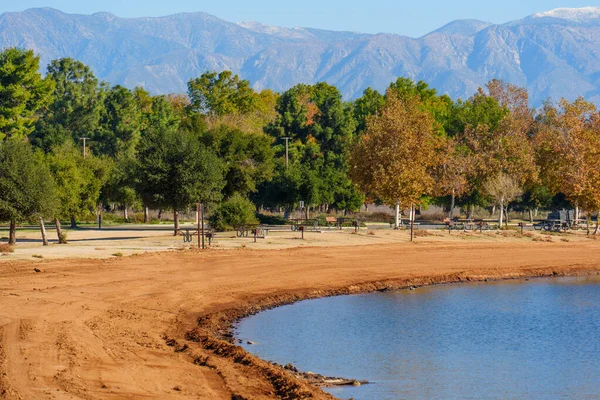 Geniş bir manzara, sakin Perris Gölü kıyı şeridini yakalar. Kumlu bir sahil, yemyeşil ağaçlar ve berrak bir gökyüzü altında karla kaplı dağlar..