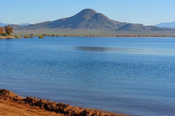 Perris Gölü 'nün sakin mavi suları parlak, bulutsuz bir gökyüzünün altında kumlu bir ön planda yer alan engebeli dağ tepelerini karşılamak için uzanır..