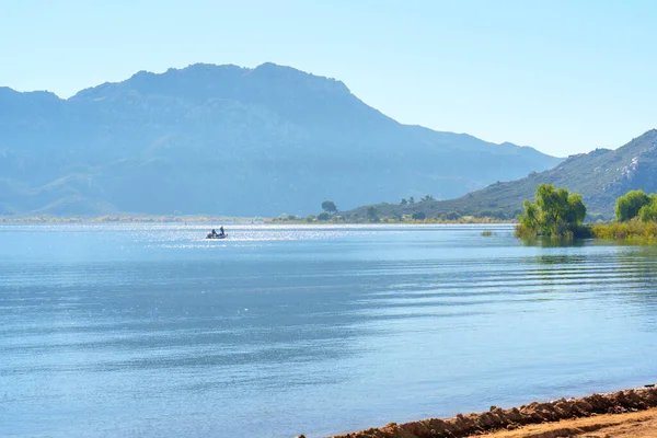 İnsanlar, Perris Gölü 'nün pırıl pırıl mavi alanında, puslu dağlar ve açık bir gökyüzü tarafından çerçevelenmiş, huzurlu bir tekne gezisinin tadını çıkarıyorlar..