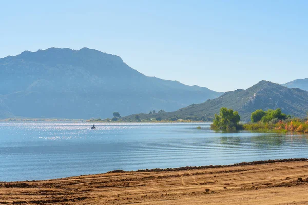 Işıldayan Perris Gölü suları berrak mavi bir gökyüzünü yansıtır, uzak puslu dağlar ve tek bir su aracı, ham, dokulu bir toprak ön planı ile.