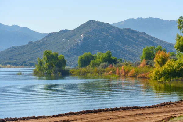 Perris Gölü suları, yeşil ve sonbahar renklerinde ağaçlarla çevrili, temiz mavi gökyüzünün altındaki uzak dağlara karşı yavaşça dalgalanır..