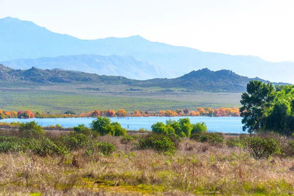 Perris Gölü 'nün sakin suları, sonbahar renklerinde ağaçlarla çevrili, kuru ön planda bitki örtüsü ve parlak, berrak bir gökyüzünün altında mavi katmanlı dağlar..