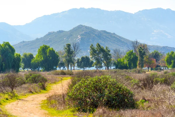 Dolambaçlı toprak yol, çeşitli yeşil ve kahverengi bitkileri aşar. Ağaçlarla ve puslu dağlarla çevrili uzak bir göle götürür..