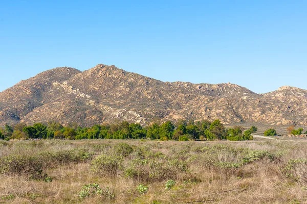 Geniş açı kuru çalı ve parlak yeşil ağaçları yakalar. Engebeli, güneşten sırılsıklam olmuş dağları mavi gökyüzünün altında..