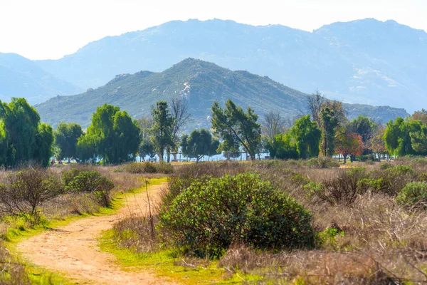 Kirli yol, bereketli ön planda çalılar ve canlı yeşil ağaçlar arasından esiyor, sakin bir göle ve uzak, sisli, mavi dağlara götürüyor..