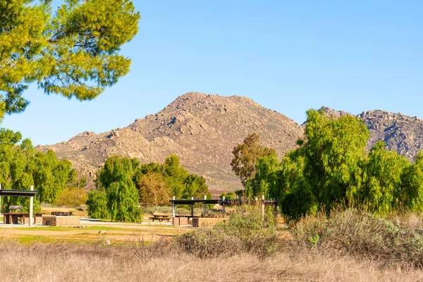 Piknik barınakları ve yemyeşil ağaçlar, Perris Gölü 'nün açık mavi gökyüzünün altında engebeli, kayalık dağlar tarafından desteklenen dinlenme alanında dururlar..