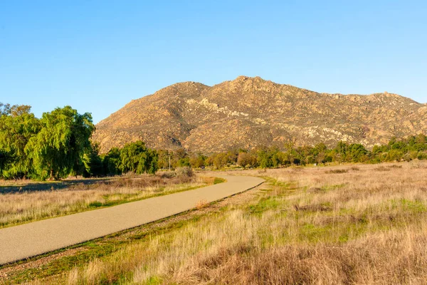 Kuru altın çimenler ve yeşil ağaçların arasından, Perris Eyaleti dinlenme bölgesinde berrak mavi bir gökyüzünün altındaki engebeli kayalık bir dağa uzanan dolambaçlı asfalt yollar. Güneşli bir günde..