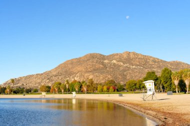 Perris Gölü 'nün sakin suları, engebeli bir dağ ve canlı ağaçlarla çevrili cankurtaran kuleleriyle dolu kumlu kıyılarla buluşuyor. Gökyüzü açık mavi, ay görünür..