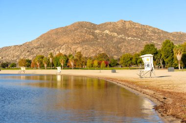 Perris Gölü Eyalet Dinlenme Alanı 'nın sakin sahnesi, sakin sular, cankurtaran standları ve engebeli bir dağın altındaki canlı sonbahar ağaçları..