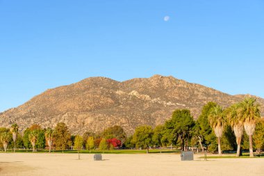 Perris Eyaleti Dinlenme Alanı 'nda çarpıcı kayalık bir dağ, canlı sonbahar ağaçları ve mavi gökyüzünde yükselen ay bulunur..