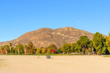 Sonbahar renkli ağaçlar, Perris Eyaleti dinlenme bölgesinde açık mavi gökyüzünün altında engebeli bir çöl dağıyla büyük bir kumlu tarlaya dizilir..