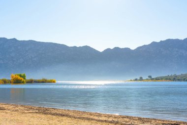 Perris Gölü 'nün berrak mavi suyu parlak güneş ışığıyla parıldıyor, önplanda kumlu bir sahil ve berrak bir gökyüzünün altında engebeli dağlar var..
