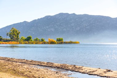 Perris Gölü 'nün sakin kıyı şeridi, kumlu kıyılar ve renkli sonbahar ağaçları, puslu uzak dağlar tarafından çerçevelenmiş parlak gündüz vakti gökyüzü altında, sakin bir doğal manzara yaratıyor..