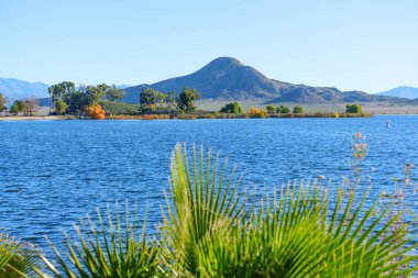 Perris Gölü 'nün durgun mavi suları berrak bir gökyüzünü yansıtır, yemyeşil ve sonbahar renkli ağaçlar tarafından çerçevelenmiş, parlak bir günde engebeli dağlarla birlikte..