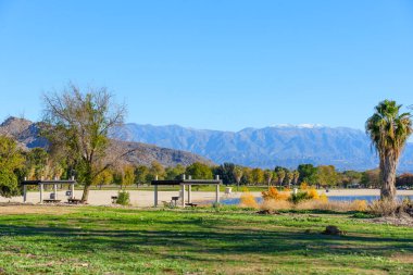 Perris Gölü Eyalet Dinlenme Alanı 'nın sakin suları, piknik barınakları ve canlı yeşillikleriyle uzak kar örtülü dağlara bakar..