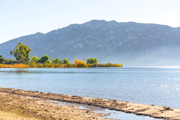 Perris Gölü 'nün sakin kıyı şeridi, kumlu kıyılar ve renkli sonbahar ağaçları, puslu uzak dağlar tarafından çerçevelenmiş parlak gündüz vakti gökyüzü altında, sakin bir doğal manzara yaratıyor..