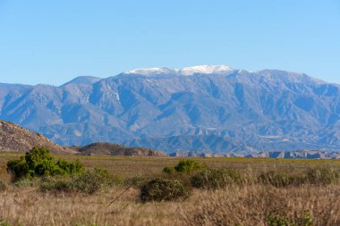 Karla kaplı zirveler, açık mavi gökyüzüne karşı görkemli bir şekilde yükselir, altın otlar ve dağınık yeşil çalılardan oluşan engin ve kuru bir manzaraya tepeden bakar..