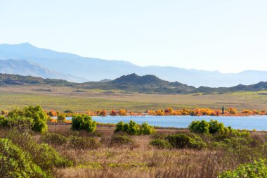 Sahne manzarası Perris Gölü 'nün mavi suyunu sunar, dalgalı dağlarla desteklenir ve sonbahar renkli ağaçlar da dahil olmak üzere çeşitli bitkilerle kaplıdır..