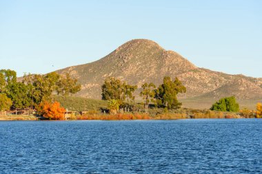 Dalgalanan mavi sular, kıyı şeridinin çeşitli yeşil ve canlı sonbahar ağaçları için görkemli kayalık dağlara karşı sakin bir ön plan sağlar..