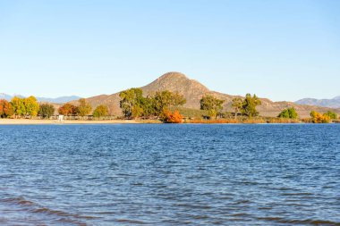 Perris Gölü 'nün sakin mavi suları gökyüzünü yansıtıyor. Arkaplanda parlak sonbahar ağaçları ve belirgin güneşli bir tepe olan kumlu bir kıyıya rastlıyor..