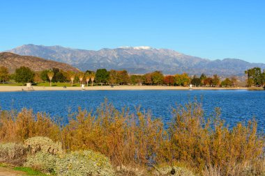 Perris Gölü 'nün berrak mavi suları parlak bir gökyüzünün altındaki görkemli karlı dağların desteklediği canlı bir sonbahar kıyı şeridini yansıtır..