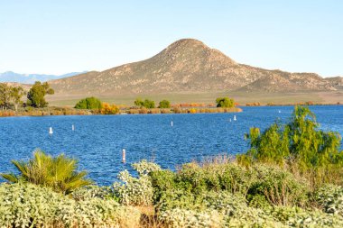 Perris Gölü 'nün engin mavi suları berrak bir gökyüzü altında dalgalanıyor. Önü çeşitli sonbahar yaprakları kaplıyor. Uzakta güneşli dağlar var..
