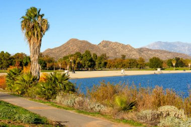 Önde gelen palmiye ağaçları Perris Gölü 'nün berrak mavi suları ve kumlu plajları, berrak bir gökyüzünün altında yükselen engebeli dağlar..