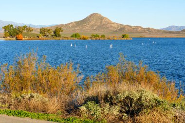 Perris Gölü 'nün ışıl ışıl mavi suları parlak bir gökyüzünün altındaki görkemli bir dağa götürür. Huzurlu ön plandaki renkli sonbahar çalılarıyla..