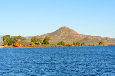 Perris Gölü 'nün mavi suları berrak bir gökyüzünün altındaki engebeli bir dağa doğru uzanır ve doğal kıyı şeridi bitki örtüsü ve beyaz şamandıralarla tamamlanır..