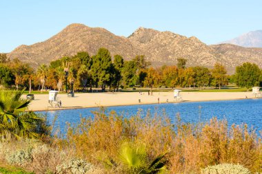 Perris Eyaleti Dinlenme Bölgesi 'ndeki sarmal patika, uzak engebeli dağlara karşı çeşitli ağaçlar, belirgin palmiyeler ve sakin göl manzaraları sergiliyor..