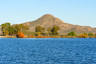 Perris Gölü 'nün mavi suları, açık ve parlak bir gökyüzü altında engebeli bir dağ tarafından desteklenen yeşil ve canlı sonbahar yapraklarıyla ağaçlarla kaplı bir kıyıya kadar uzanır..
