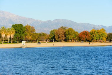 Perris Gölü 'nün mavi suları temiz gökyüzünü yansıtır. Beyaz cankurtaran kulesi, insanlar, çeşitli ağaçlar ve uzak dağlarla kumlu bir sahile dönüşür..