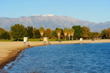 Sakin mavi göl suyu, açık mavi gökyüzü altında yeşil ağaçlar ve uzak karlı dağlar tarafından desteklenen üç beyaz cankurtaran standının olduğu kumlu bir plajı kaplar..
