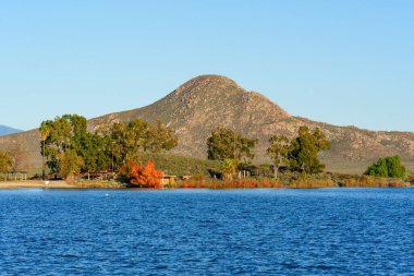 Perris Gölü 'nün mavi suları berrak bir gökyüzünü yansıtırken, sonbahar renkli ağaçlar arka planda belirgin bir dağla kıyı boyunca uzanır..