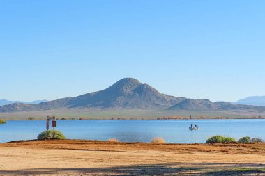 Parris, California - 7 Aralık 2025: Perris Gölü 'nün mavi sularındaki bir tekneden iki kişi, belirgin bir dağ ve berrak gökyüzü ile, barışçıl bir açık hava eğlencesi tasvir ediyor..