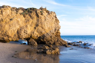 Altın güneş ışığı, El Matador Eyalet Sahili 'ndeki görkemli bir doğal kaya kemerini güzelce aydınlatıyor. Köpüklü okyanus dalgaları, kumlu sahili hafifçe okşuyor..
