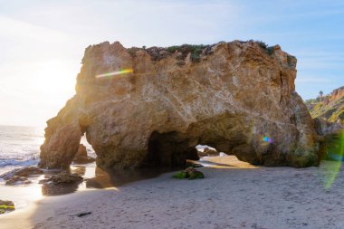 Parlak altın ışık, El Matador Eyalet Sahili 'nde belirgin bir kaya kemerini aydınlatıyor. Engebeli oluşumları vurguluyor ve okyanus kıyısında ıslak kum parlıyor..