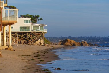 Sağlam ayaklıklardaki kıyı evleri, açık mavi gökyüzü altında kumlu bir sahil, yumuşak okyanus dalgaları ve belirgin kaya oluşumlarının yer aldığı El Matador State Beach 'e bakar..