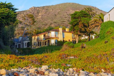 Kırmızı çatılı ve balkonlu sarı sıva tarzı villa Malibu 'daki El Matador Plajı' ndaki yeşil bir yamaçtan kayalık bir plaja bakıyor..