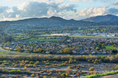 Geniş Riverside, California yerleşim alanları ve canlı yeşil alanlar güzel, parçalı bulutlu bir gökyüzü altında engebeli dağlara doğru uzanır..