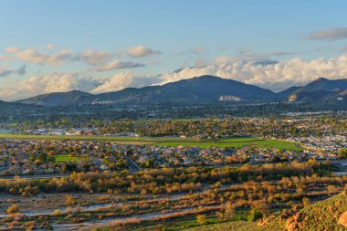 Altın saat ışığı uzak dağlar ve bulutlu gökyüzü tarafından çerçevelenmiş vadideki yerleşim bölgelerini, yeşil tarlaları ve kuru nehir yatağını aydınlatır..