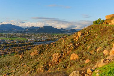 Geniş bir şehre doğru geniş kayalarla süslenmiş engebeli, çalılarla kaplı yamaçlar ve engin, parçalı bulutlu gökyüzünün altındaki uzak dağlar..