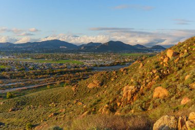 Geniş manzaralı manzara, Riverside, California 'nın engin kentsel yayılımını yakalar. Yumuşak, altın saat gökyüzü altında görkemli dağlara yaslanır..
