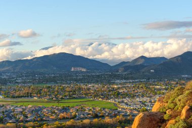 Riverside, California 'ya bakış açısıyla, yeşil manzaralar arasında şehir sergileniyor, etkileyici dağlar ve pofuduk bulutlar manzaralı bir zemin oluşturuyor..