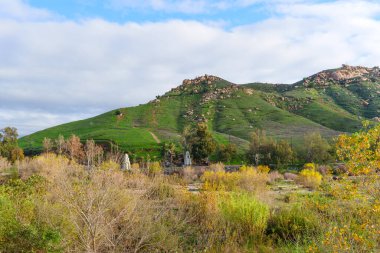 Rubidoux Dağı 'nın canlı yeşil yamaçları dinamik gökyüzü altında yükselir, sonbahar önü çalıları ve iki farklı anıtla çelişir..