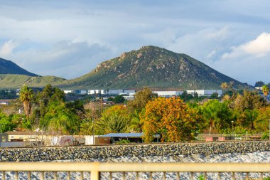 Görkemli Rubidoux Dağı parçalı bulutlu bir gökyüzünün altında yükseliyor, endüstriyel binaları, çeşitli yeşillikleri ve Riverside, California 'da kayalık bir seti denetliyor..