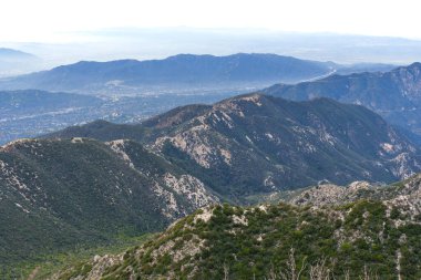 Koyu yeşil, şapel kaplı San Gabriel Dağları 'nın geniş Los Angeles havzasına bakan yüksek açılı manzarası kalın bir sis tabakasıyla örtülmüş..