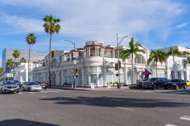 Beverly Hills, California - 21 Ocak 2026: Güneşli bir günde dünyanın en ünlü Rodeo Drive caddesi köşesinde park edilmiş palmiye ağaçları ve lüks araçlarla lüks bir perakende mimarisi.