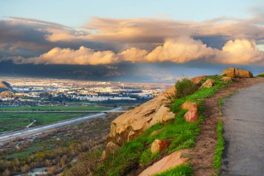 Riverside, Kaliforniya 'daki Rubidoux Dağı' ndan panoramik manzara, sarmal bir yol gösteriyor, yemyeşil yamaç ve dramatik günbatımı bulutları altında kentsel vadi..