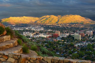 Riverside şehrinin ufuk çizgisi ve Box Springs Dağı, Rubidoux Dağı 'nın basamaklarından görülen dramatik fırtına bulutlarının altında altın saat güneş ışığıyla aydınlandı..